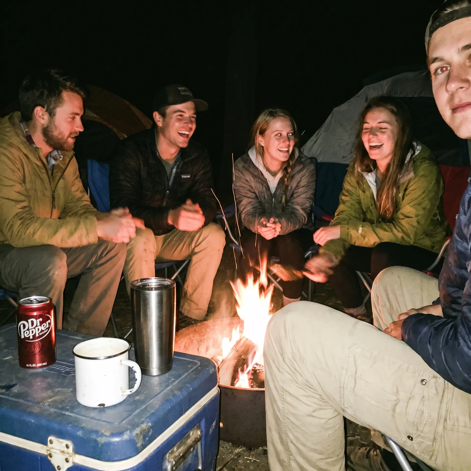 Friends laughing around a campfire at night beside their tents